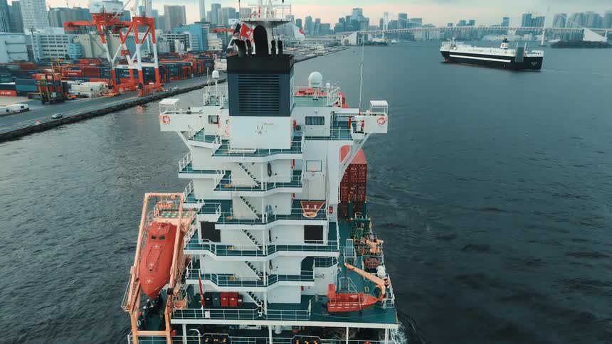 An aerial view shows a massive container ship sailing through a harbor towards a modern city skyline with a bridge visible in the distance the ships deck is stacked high with numerous red cargo containers ready for transport014