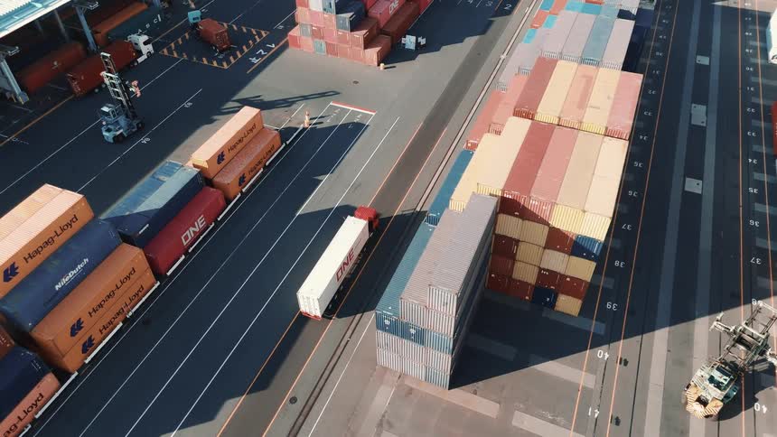This drone footage shows an aerial view of a busy industrial port the video focuses on a forklift that is carrying and maneuvering a large shipping container between stacks of other containers