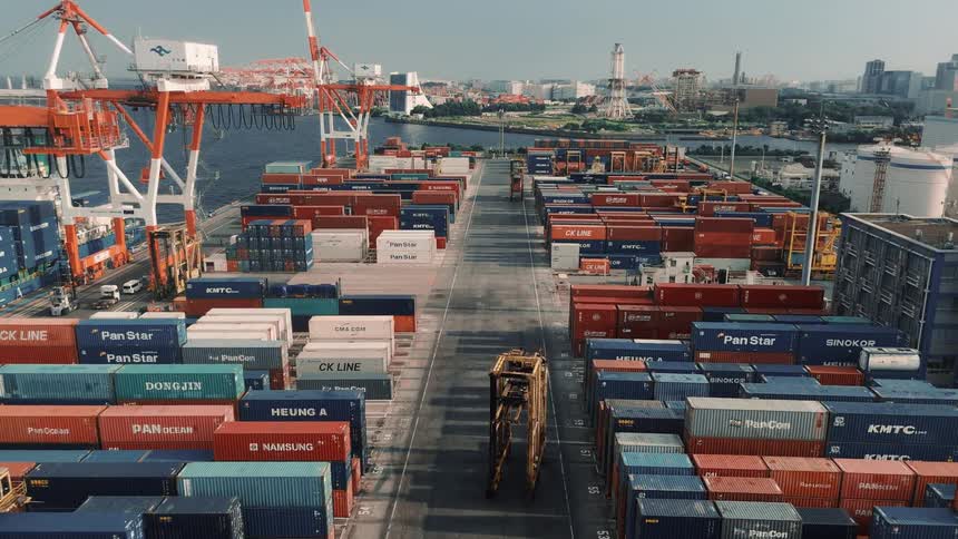 A busy seaport at midday packed with towering stacks of colorful containers massive gantry cranes loom overhead while loaders move swiftly across the yard lifting and transporting containers under the bright sun