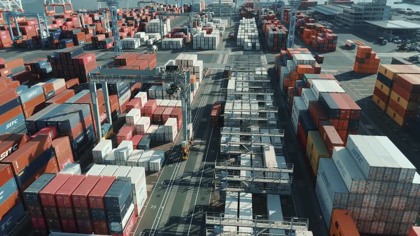 This drone footage captures an aerial view of a busy container port the video showcases large cranes moving and stacking a vast number of shipping containers in various colors with the sea visible in the background