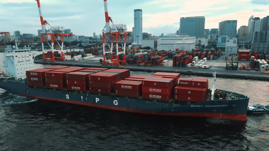 An aerial view shows a cargo ship loaded with bright red containers sailing away from a busy harbor in the background large cranes stand ready on the dock with a modern city skyline stretching out behind them