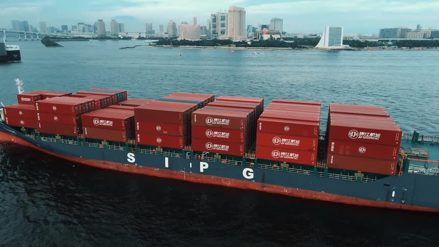 A large cargo ship marked sipg sails near a city shoreline stacked high with rows of red shipping containers