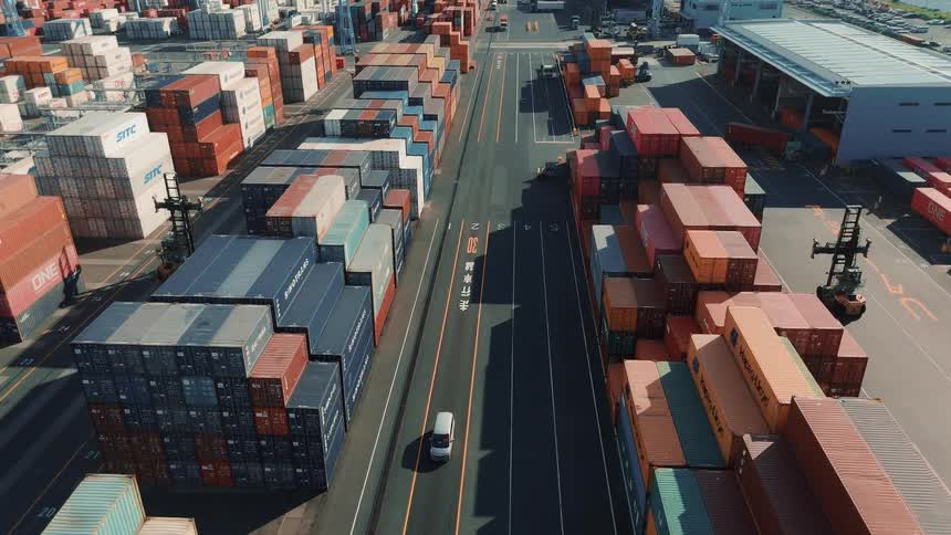 A seaport is filled with stacked cargo containers with cars and trucks driving along a tarred road that winds through the containers at midday