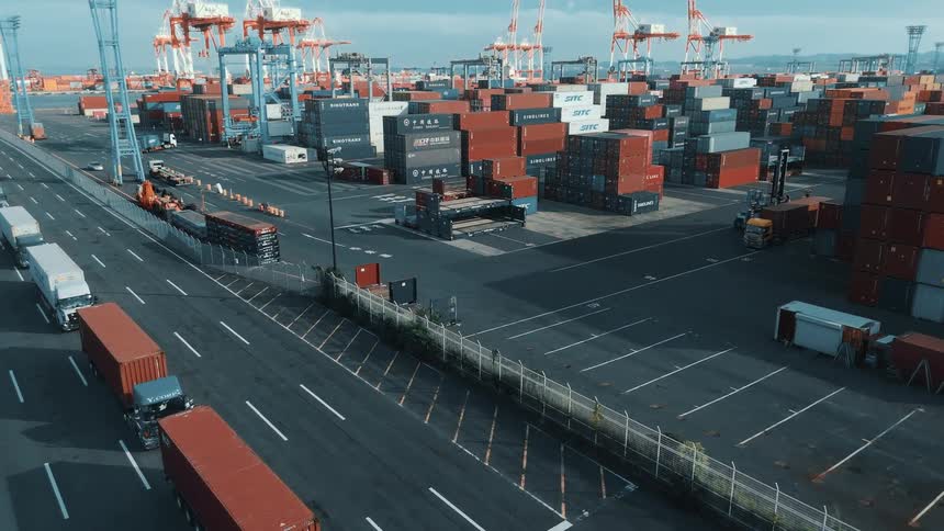 A busy seaport at midday packed with towering stacks of colorful cargo containers as trucks loaded with container freight drive in and out along the paved lanes a line of massive cranes stands tall against the bright sky under the bright sun