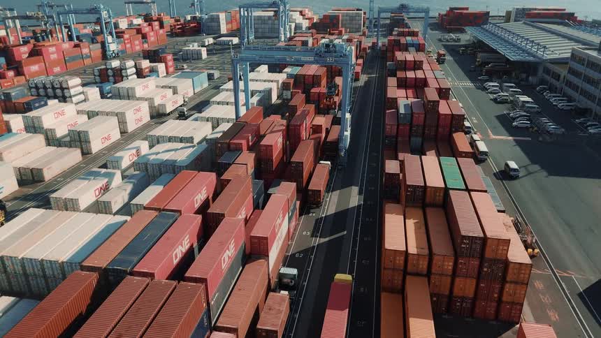 A sea port filled with stacked shipping containers with trucks driving in and out of the port under a bright sunny sky