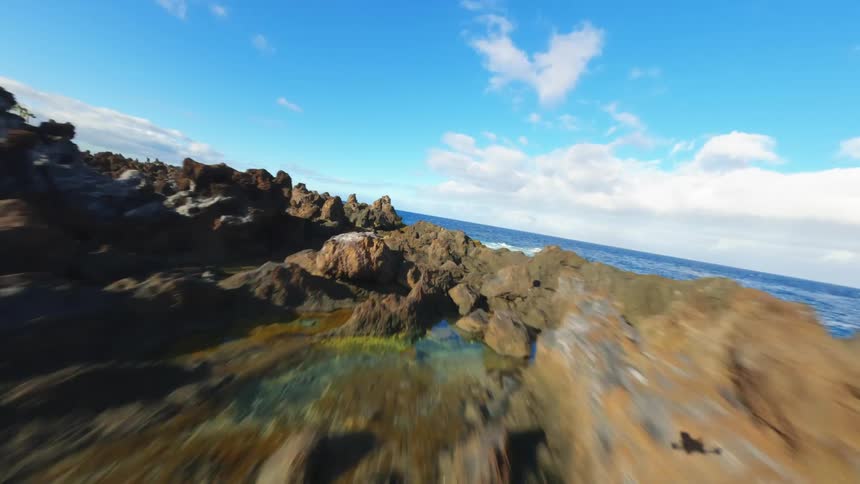 This aerial footage captures a drone flying over a rocky coastline with natural sea pools the camera follows the rugged volcanic rock formations jutting out into the deep blue ocean