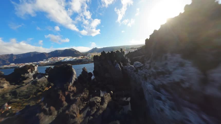 This is a drone video filmed at a rocky coastline with a clear blue sky and ocean the drone flies over a natural tide pool and then swoops out over the ocean revealing a distant town and mountains along the coast