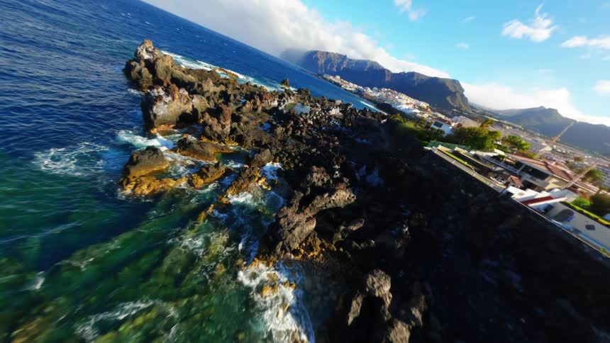 A rocky coastline with rugged uneven terrain stretches along the shore under the bright midday sun with waves crashing against the rocks beyond the coastline the distant city skyline rises against the clear horizon