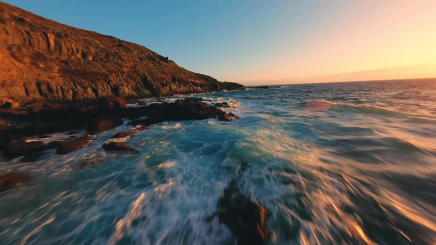 Golden sunlight illuminates rugged coastal cliffs as waves crash against the rocky shoreline