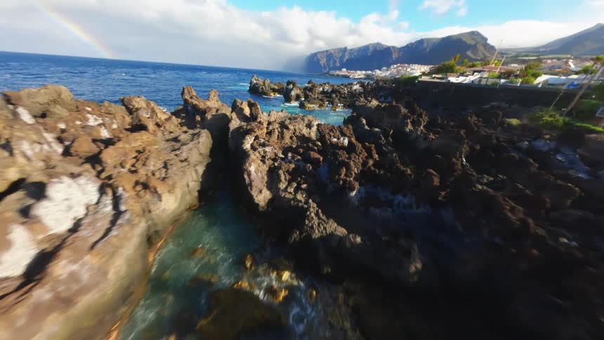 This is a highangle drone shot of a rocky coastline the drone moves over jagged black rock formations revealing tide pools with calm clear water and the vast blue ocean beyond