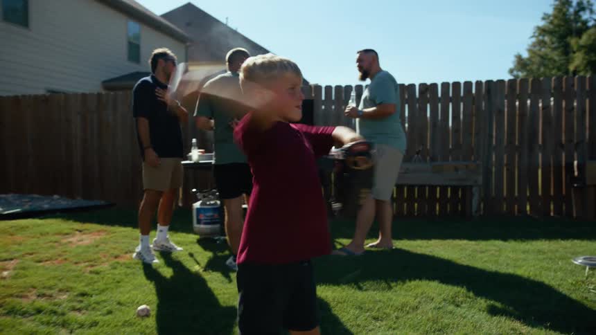 A boy with a glove stands in a yard while a group of men talks behind him