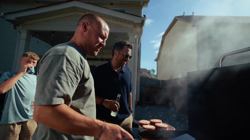 People stand around watching a man grill hamburgers on a slightly cloudy day