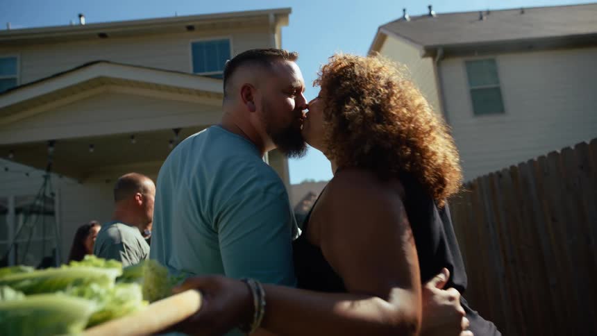 A man and a woman hold platters of food and kiss in a yard while there other people in the background
