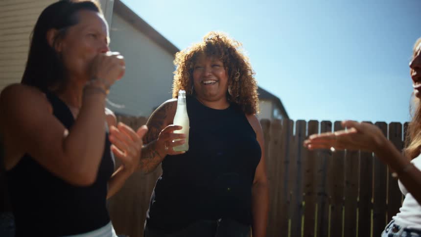 Three women are laughing and talking together outdoors with a wooden fence and a house in the background the woman in the middle who has curly hair is holding a bottle and appears to be telling a story