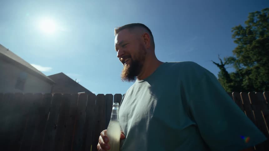 Man with a long beard wearing a blue tshirt standing beside his backyard fence next to a smoking grill holding a bottle of drink in his hand at midday