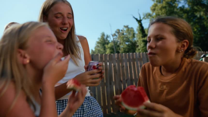 Three girls stand outside in a backyard laughing and eating watermelon on a sunny day one of them holds a drink can as they smile and talk to each other