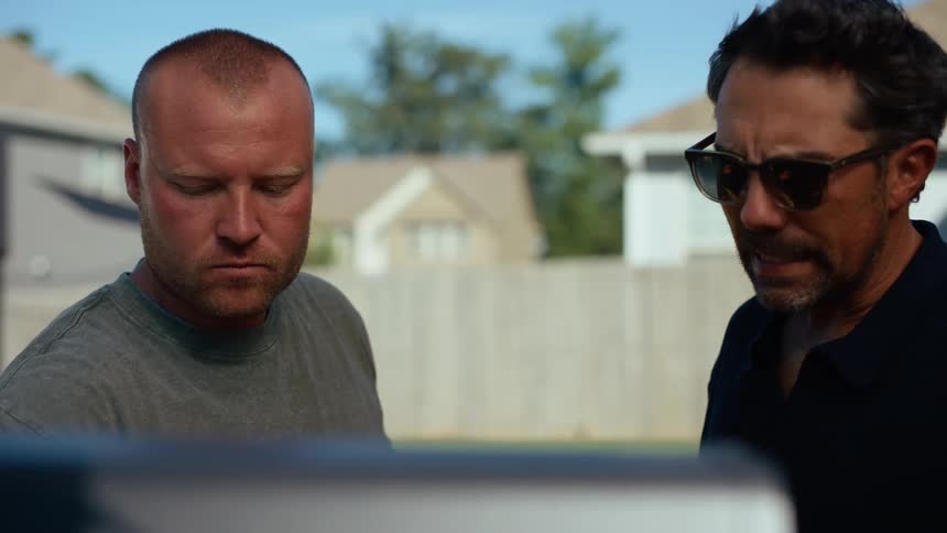 Two men are seen outdoors with one drinking from a bottle and the other looking down