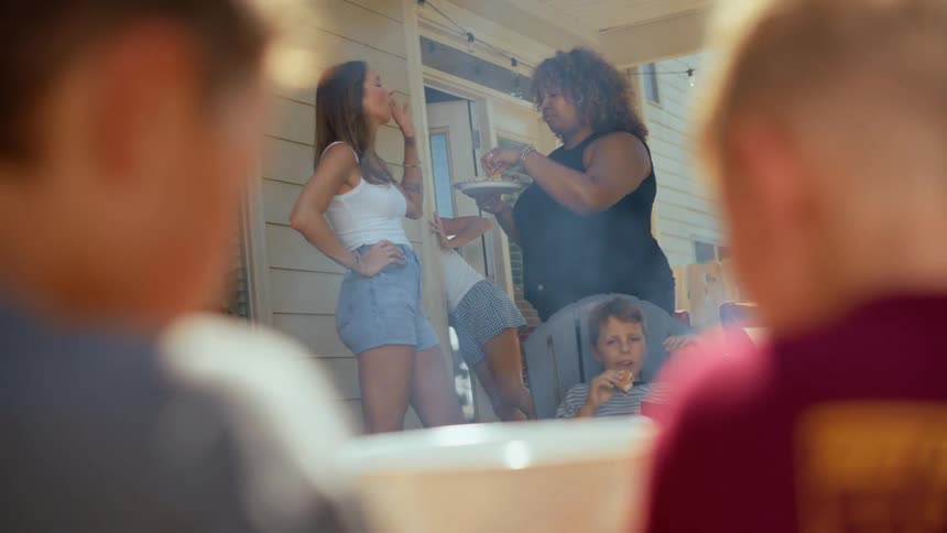 Two women chat and eat while standing on a porch as children sit nearby enjoying food at what looks like a backyard gathering