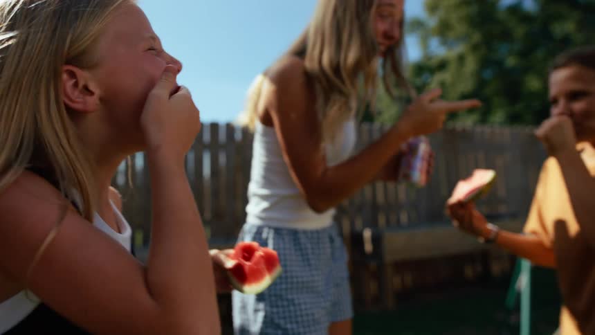 Three kids laugh and snack on watermelon slices in a sunny backyard