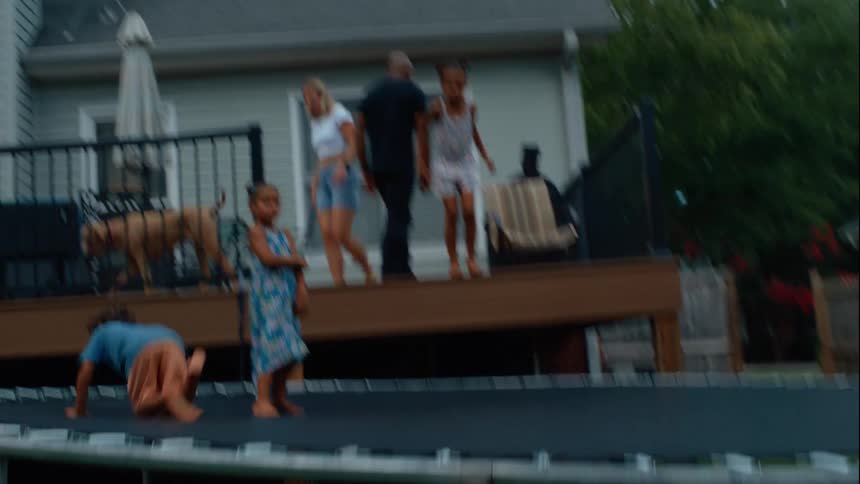 Three kids jump together on a trampoline in a sunny backyard at midday while their parents stroll across the porch with a dog nearby enjoying the warm bright day