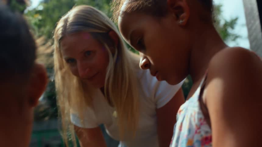 A blonde woman in a white tshirt and two children are in a garden with the children in the foreground the woman is showing a small plant to the children and all three are looking down at it