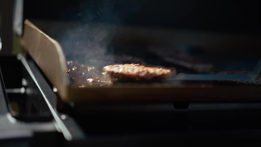 A video shows several hamburger patties sizzling on a flattop grill a person uses a spatula to flip one of the patties