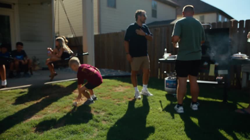A boy with a baseball glove plays in the yard while adults gather around a smoking grill in the background