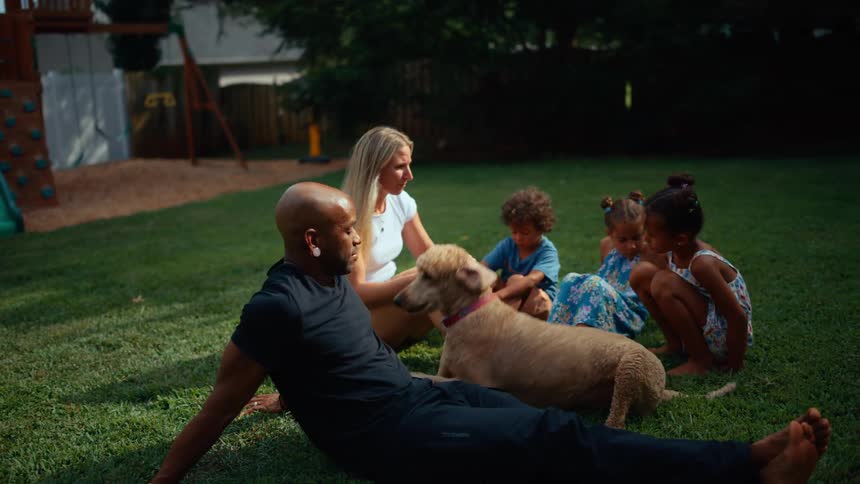 A family relaxes on the grass as children play and a dog nuzzles a man while a woman looks on