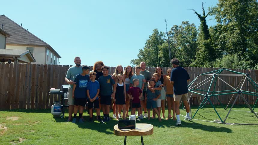 A large family is posing for a group photo in a sunny backyard with a man setting up a camera on a small table in the foreground