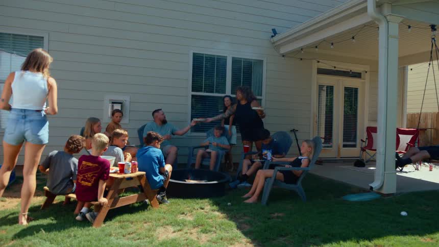 A diverse group of family and friends are gathered in a backyard for a barbecue the adults are holding plates of food and talking while the children are sitting at a picnic table and playing on a swing