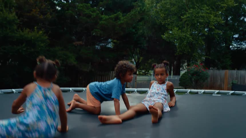 A young girl jumps on a trampoline in a sunny backyard at midday while a boy and another girl sit on the trampoline watching her the trampoline bounces from the impact of her jump surrounded by the bright open yard