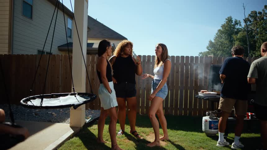 A lively backyard scene at midday with friends and siblings gathered together two kids play on a swing while a group of boys stand around a grill drinking and cooking food off to one corner the girls chat and laugh under the sun