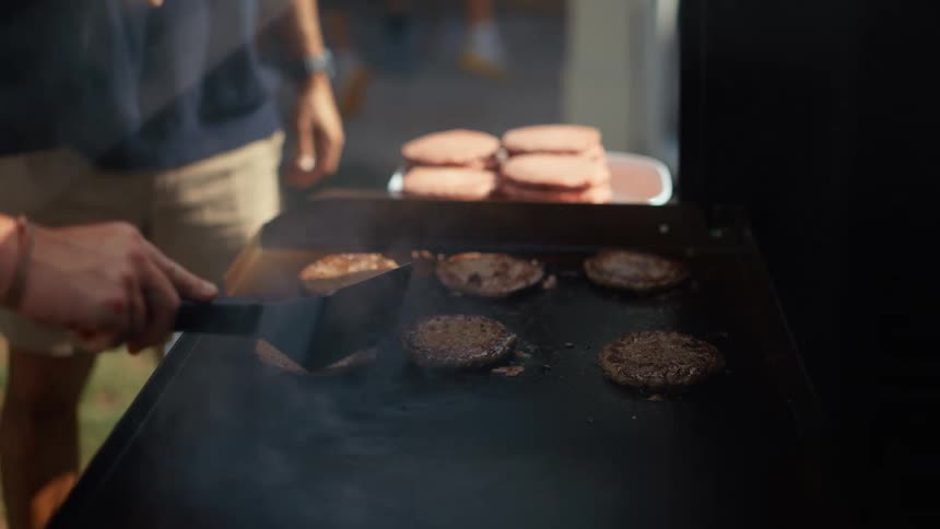 A man uses a spatula to flip food on a grill while another man stands nearby