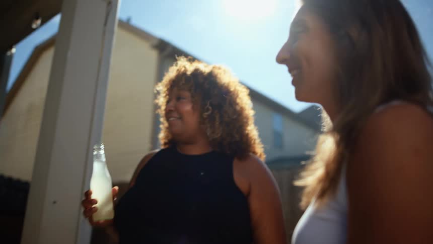 Two people stand outside on a sunny day holding glass bottles and smiling one person with long brown hair looks at a person with curly hair and they are talking to each other