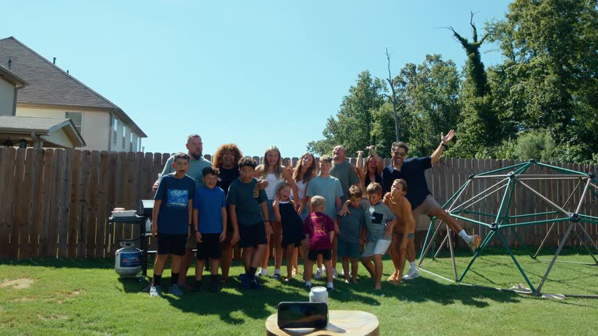 A man uses his smartphone to take a group photo of a multigenerational family including men women and children gathered in a sunny backyard with a wooden fence and playground equipment visible in the background