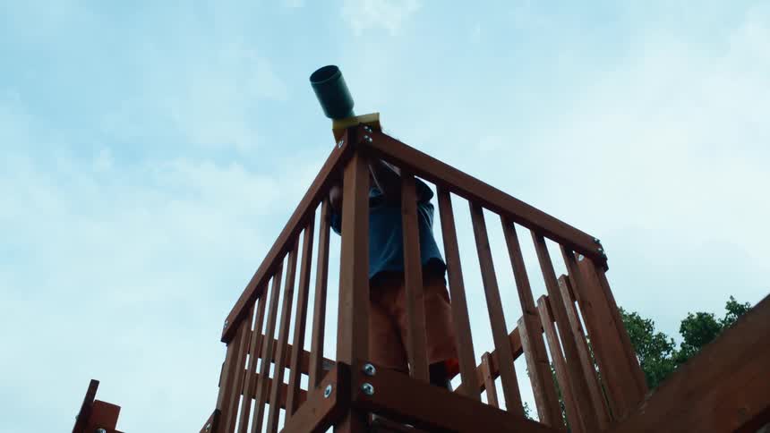 A child stands on a wooden platform in a play area at midday staring into a pair of binoculars