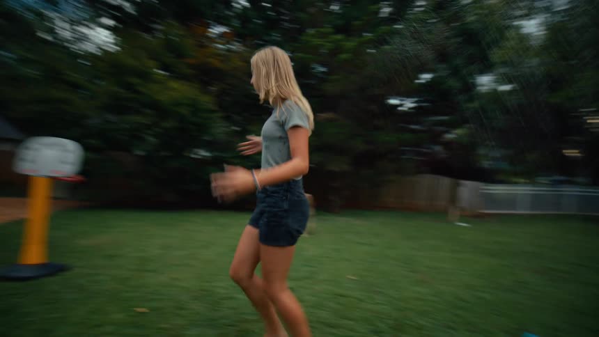 A happy group of barefoot children and a young woman are running and laughing while playing with a sprinkler on a lush green lawn in a backyard