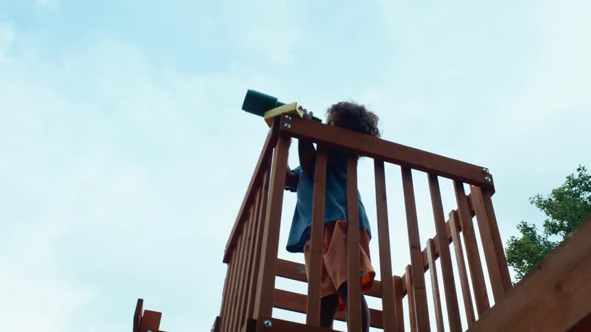 A young child stands at the top of a wooden playset looking through a toy telescope they are wearing a blue shirt and orange shorts and appear to be looking for something in the distance