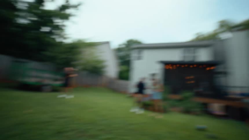 A man wearing a black shirt is throwing a basketball in a backyard during a family party with other people and a playground visible in the background