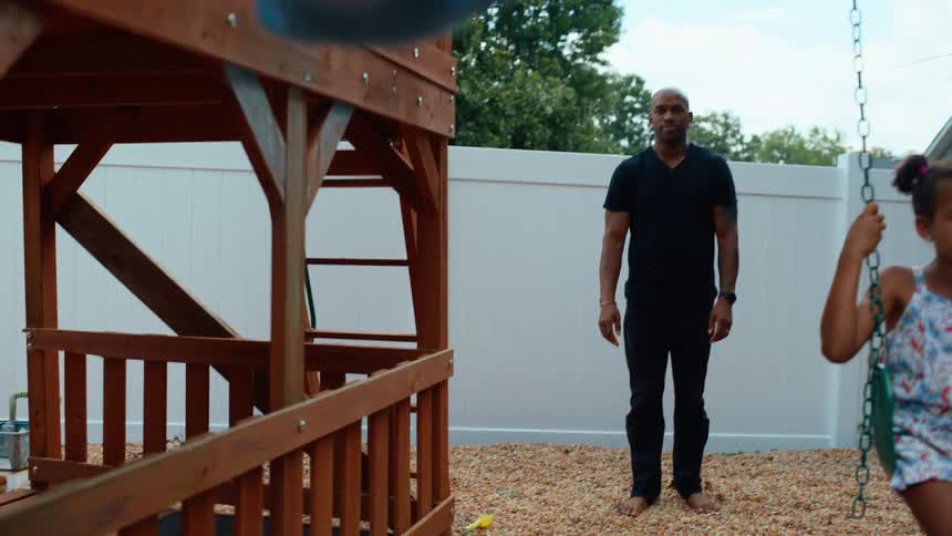 A man watches over two children swinging on a backyard playset one smiling as they swing forward and the other looking thoughtful