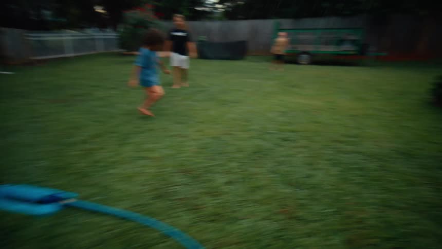 Male toddler playfully running over a water sprinkler surrounded by other children and a teenager on a lawn outdoors during day time
