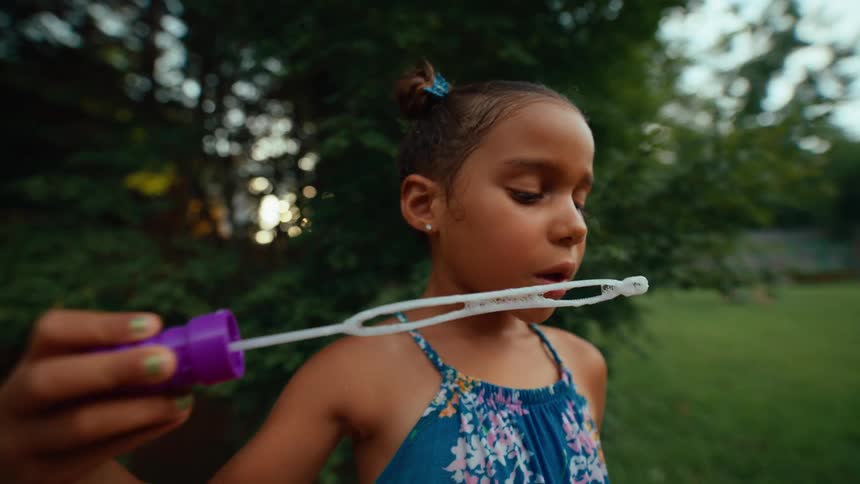 A young girl in a blue floral dress is blowing bubbles in a garden at sunset