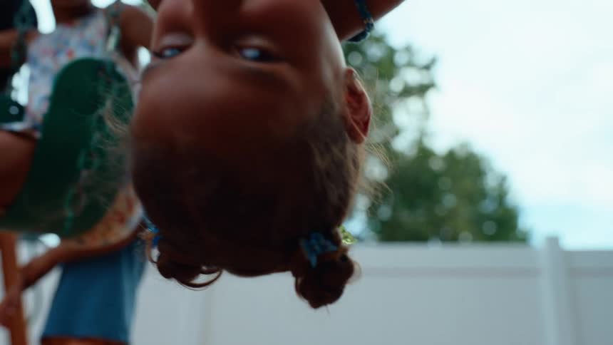 A happy young girl with her hair in braids is hanging upside down from a swing set smiling directly at the camera with other children playing in the background