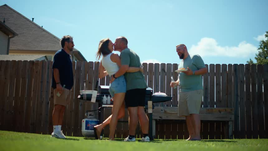 A man and woman hug and kiss in a backyard with a man and woman in the background the man and woman are smiling and laughing as they kiss