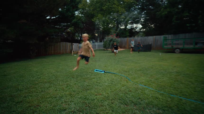 Kids run and jump over water spraying from a blue hose on a backyard lawn at midday while parents friends and family stand to the side watching the event