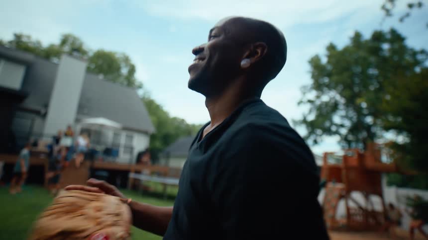 In the foreground a black man happily throws a baseball in the background a group of people are having fun together in a backyard