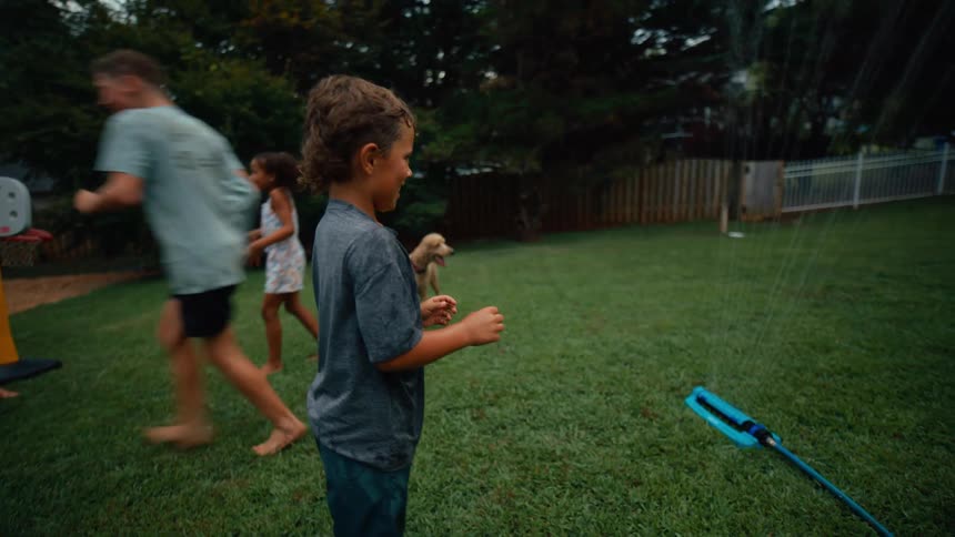 A group of children run and jump over a sprinkler in a yard
