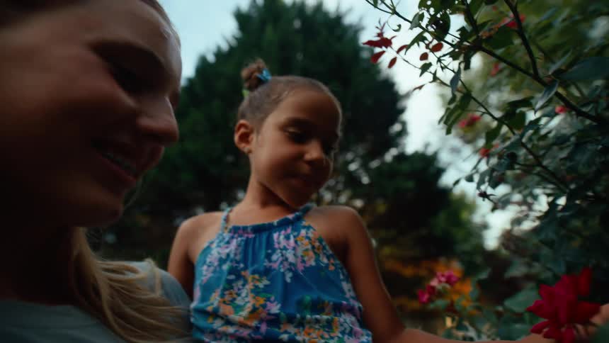 A smiling woman holds a young girl in a floral dress close to blooming roses in a garden