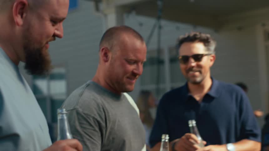 Three men smile and chat closely enjoying themselves at an outdoor gathering drinking from bottles