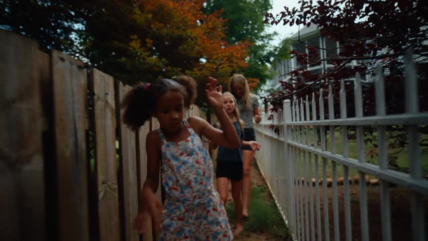 Four girls walk down a walkway between a wooden fence and a metal fence with a house visible in the background as one of the girls at the front waves her arm